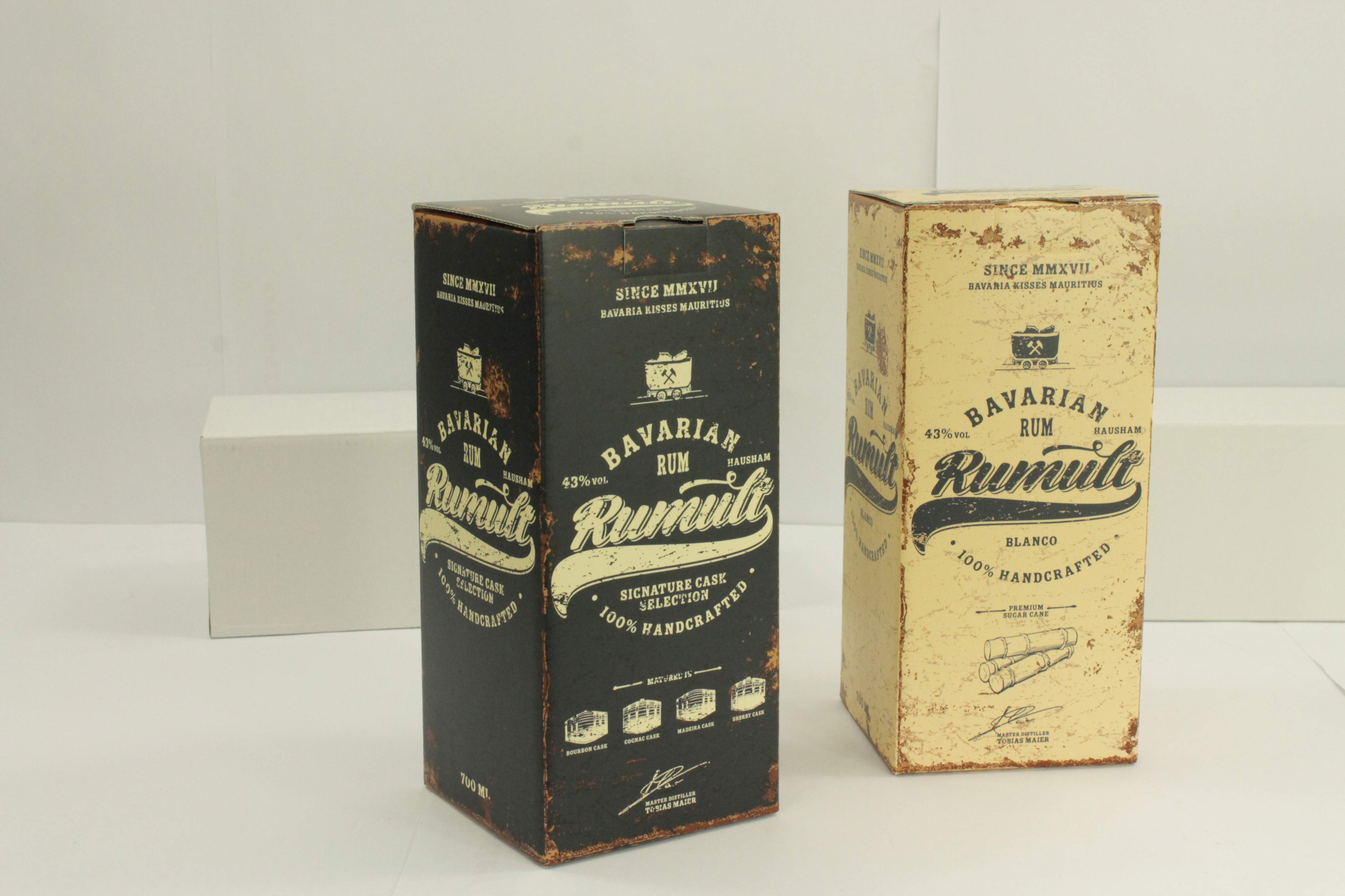 Zwei Verpackungen von bayerischem Rum (Bavarian Rum) in unterschiedlichen Designs. Die eine Faltschachtel ist dunkelblau / schwarz mit gelber Schrift und die andere ist gelb mit dunkeler Schrift