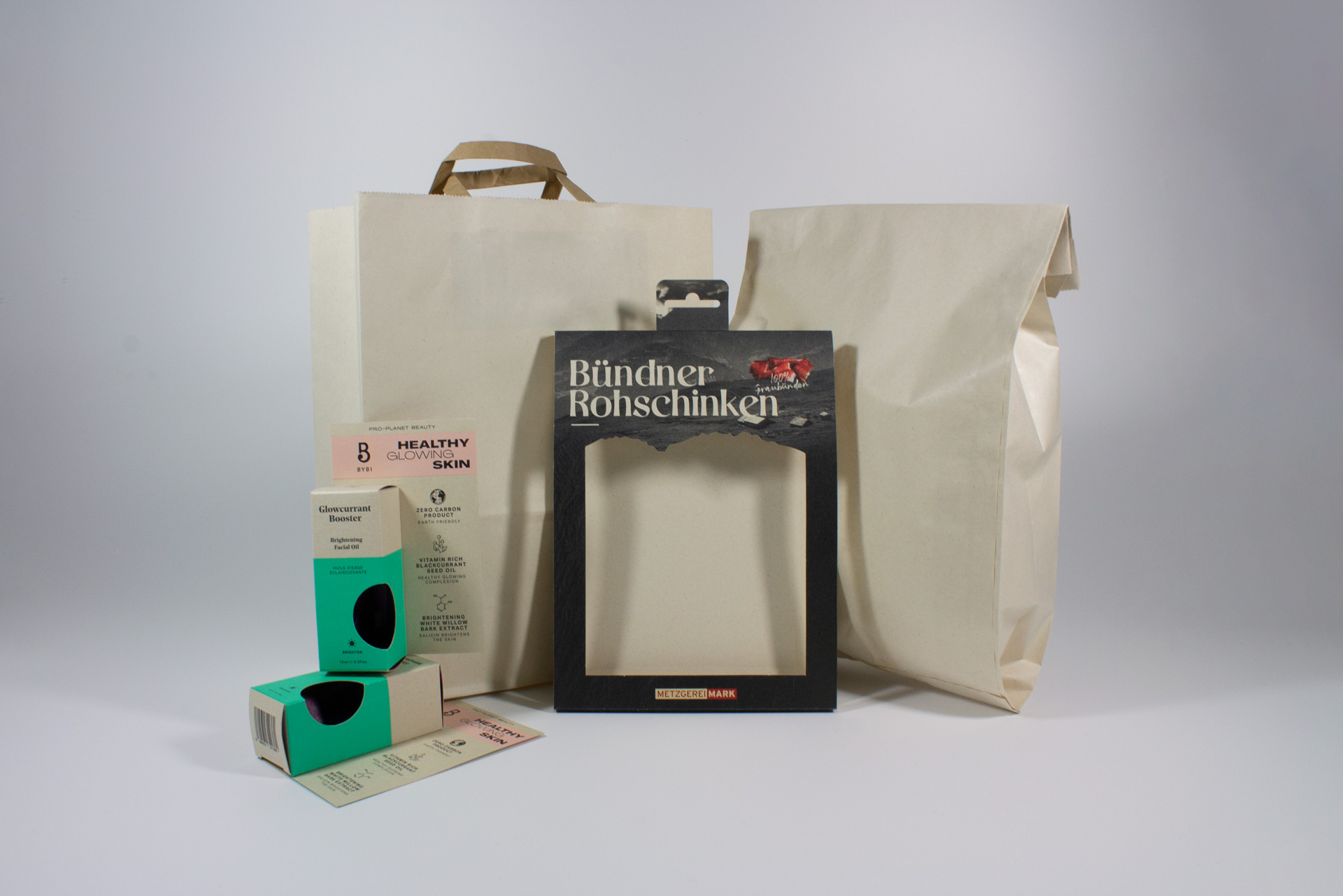 Verschiedene nachhaltige Verpackungen aus Zuckerrohrpapier: Eine Faltschachtel mit Sichtfenster, eine Banderole mit schwarzem Druck, eine naturfarbene Tragetasche und eine naturfarbene Bäckertüte