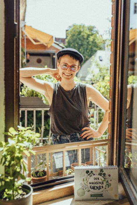 Manuela steht auf einem Balkon hinter einem geöffneten Fenster. Vor ihr steht "Der Wachsende Kalender"